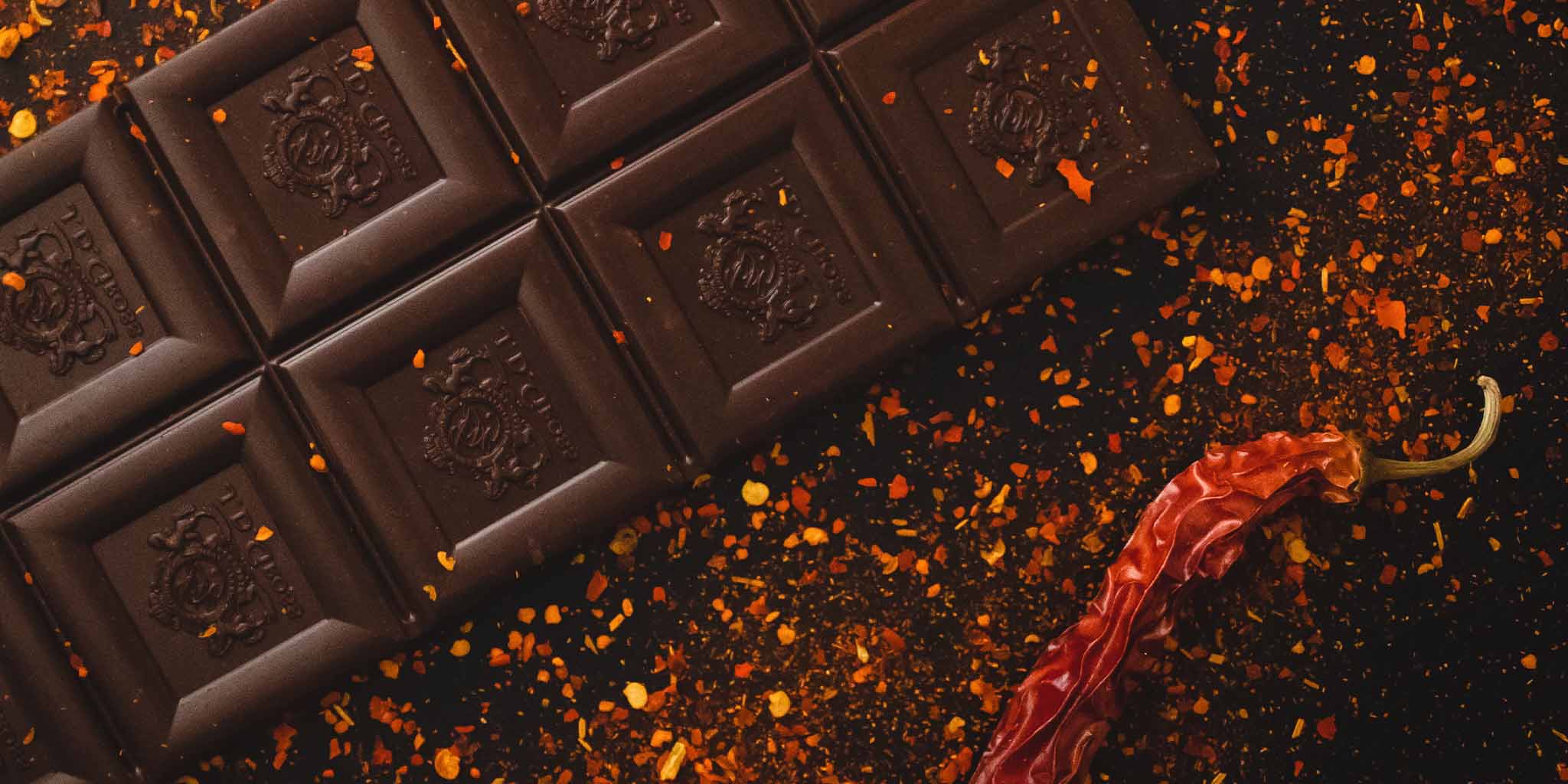 Closeup of a luxurious bar of dark chocolate sprinkled with dried chili flakes; a dried red chili sits casually beside the bar of chocolate
