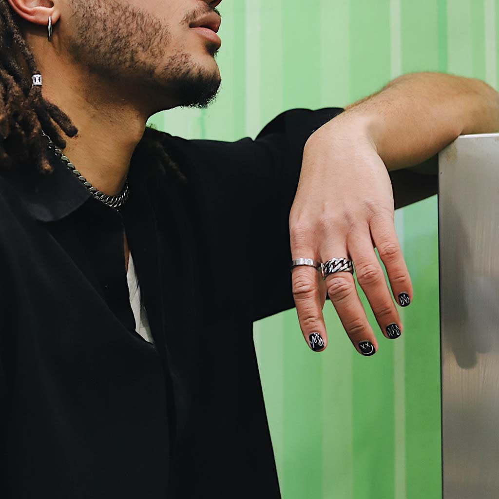 An attractive person with a short beard and dreadlocks rests an elbow on a metallic platform, allowing a hand to dangle casually; rings and fingernail polish accentuate the beauty of the figure.