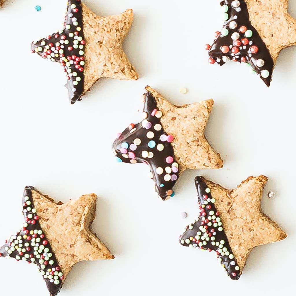 Five star-shaped cookies are each half-dipped in dark chocolate and sprinkles; they are placed against a white background with a few crumbs casually left in view.