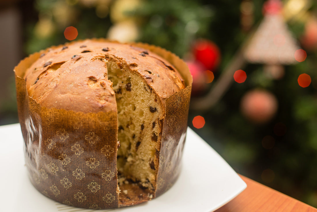A loaf of panettone sits on a plate; a slice has been cut out of the loaf, giving a glimpse of the candied citrus fruits that have been baked into the bread. An out-of-focus Christmas tree is in the background. Image from Wikipedia.