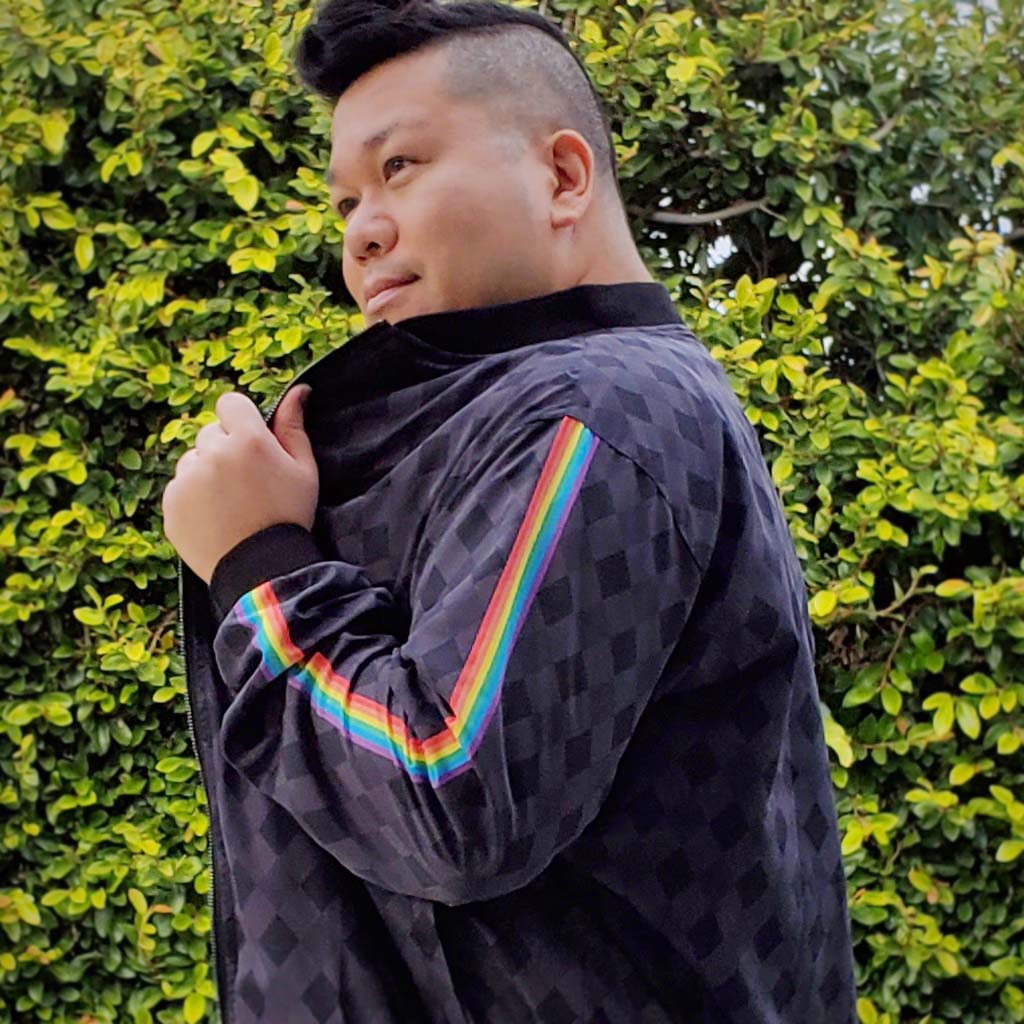 Person wears a jacket in the Rainbow Black Truffle pattern by My Friend Ren; in the background are lush green vines.