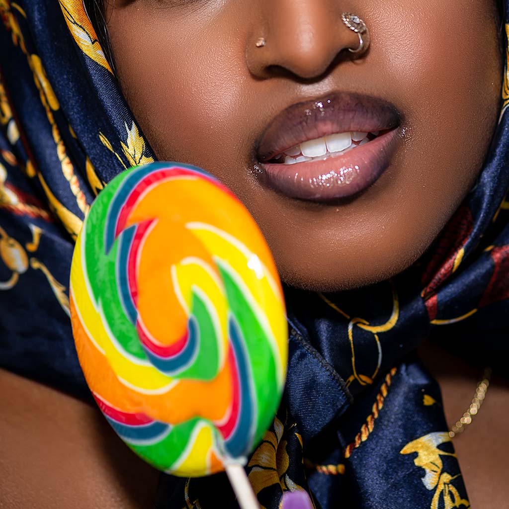 Closeup photo of a gorgeous dark-skinned woman holding a giant colourfully swirled lollipop near her perfectly glossed lips