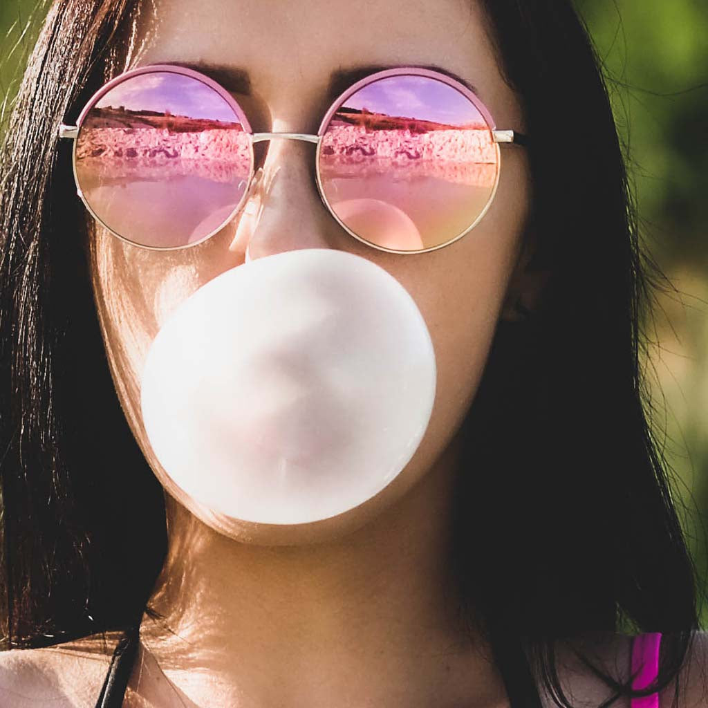 Closeup shot a long-haired person wearing reflective pink sunglasses blowing a large bubblegum bubble.