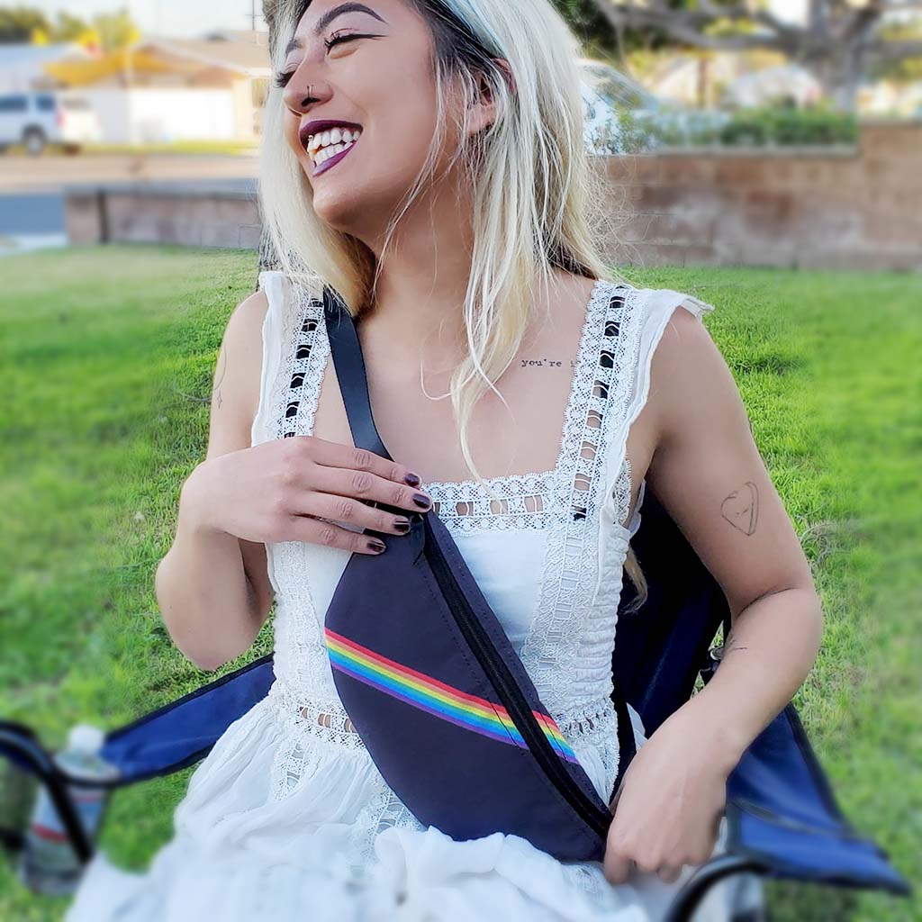 A beautiful woman of color sits in a lawn chair outdoors; she wears a hip pack as a cross-body bag; her smile is bright and magnetic. Featuring the Slim Rainbow design by My Friend Ren.
