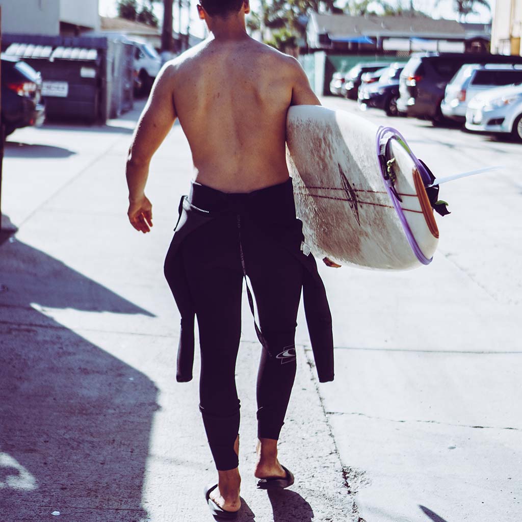 A half-nude surfer carries a surfboard while his wetsuit is pulled down to his waist. In the background is a blurred parking lot with cars.