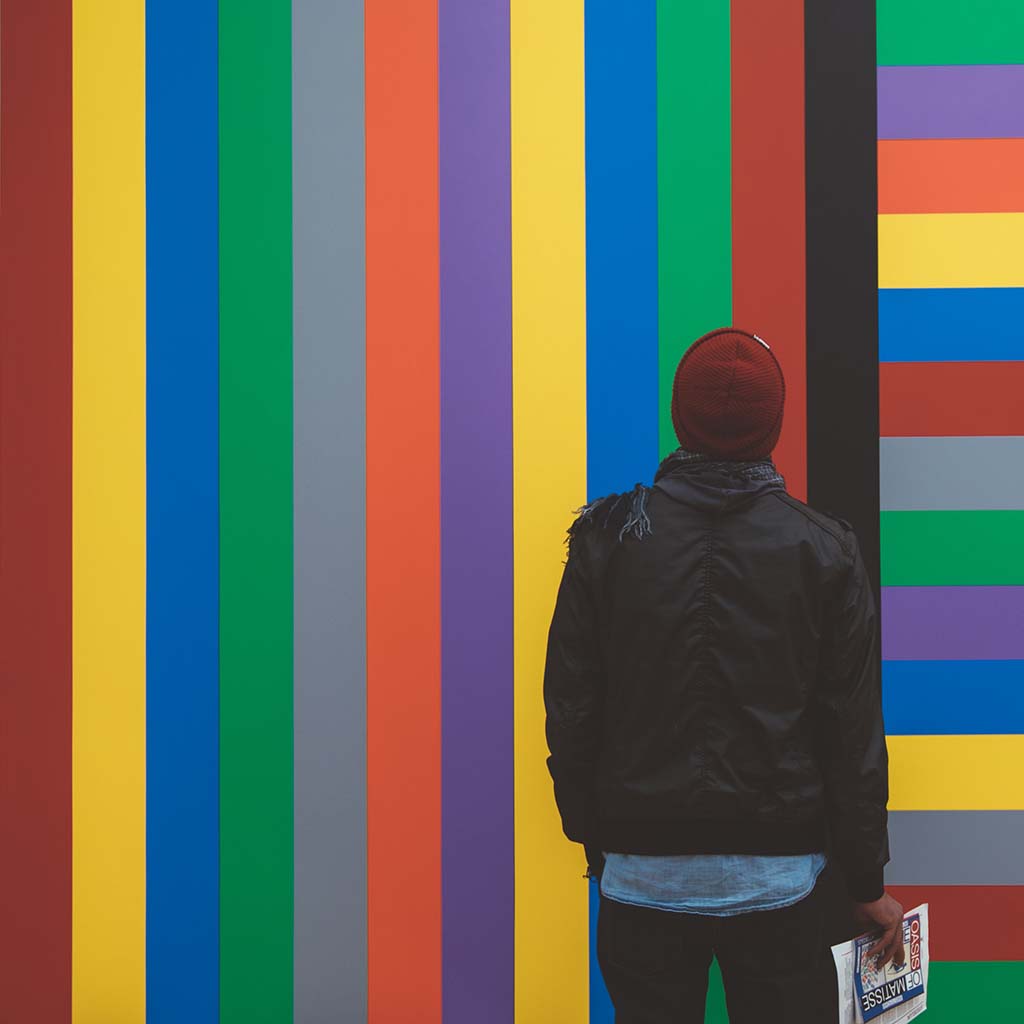 A male figure stands with his back to us as he faces a wall painted with colourful strips, most of which are vertical; at the very right of the photo, there is a hint of horizontal stripes of colour.