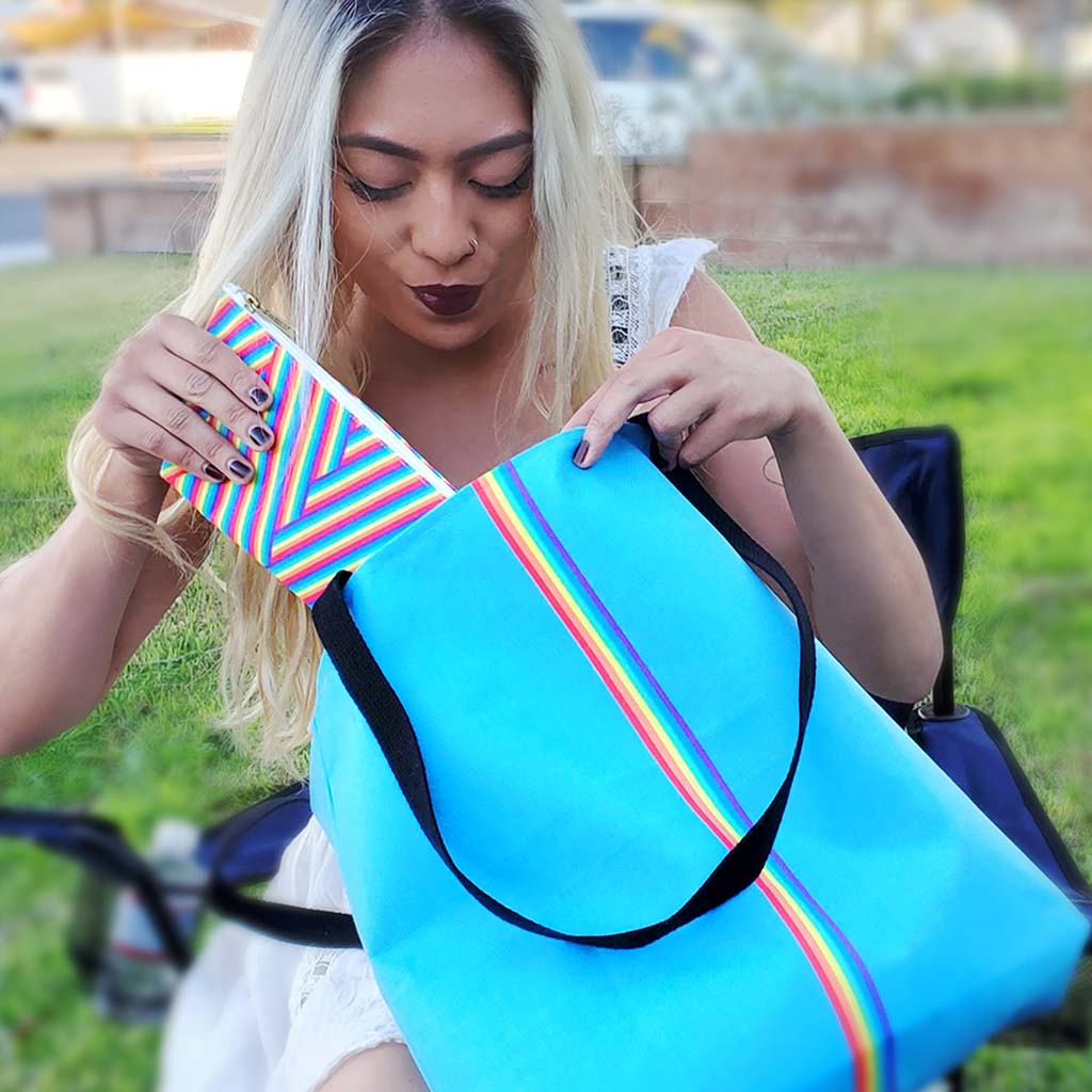 A seated woman holds a zippered pouch in the Rainbow Herringbone design and a medium sized tote bag in the Robin's Egg Slim Rainbow design, both by My Friend Ren.
