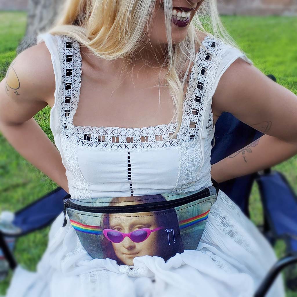 A beautiful woman of colour wears a white dress and sits on a lawn chair outdoors; she is buckling a hip pack around her waist. Featuring the Rainbow Mona Lisa design by My Friend Ren.